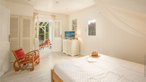 A bedroom with a bed, a television, a small table with a lamp, and a window with a view of greenery. - Granada (Le Diamant)