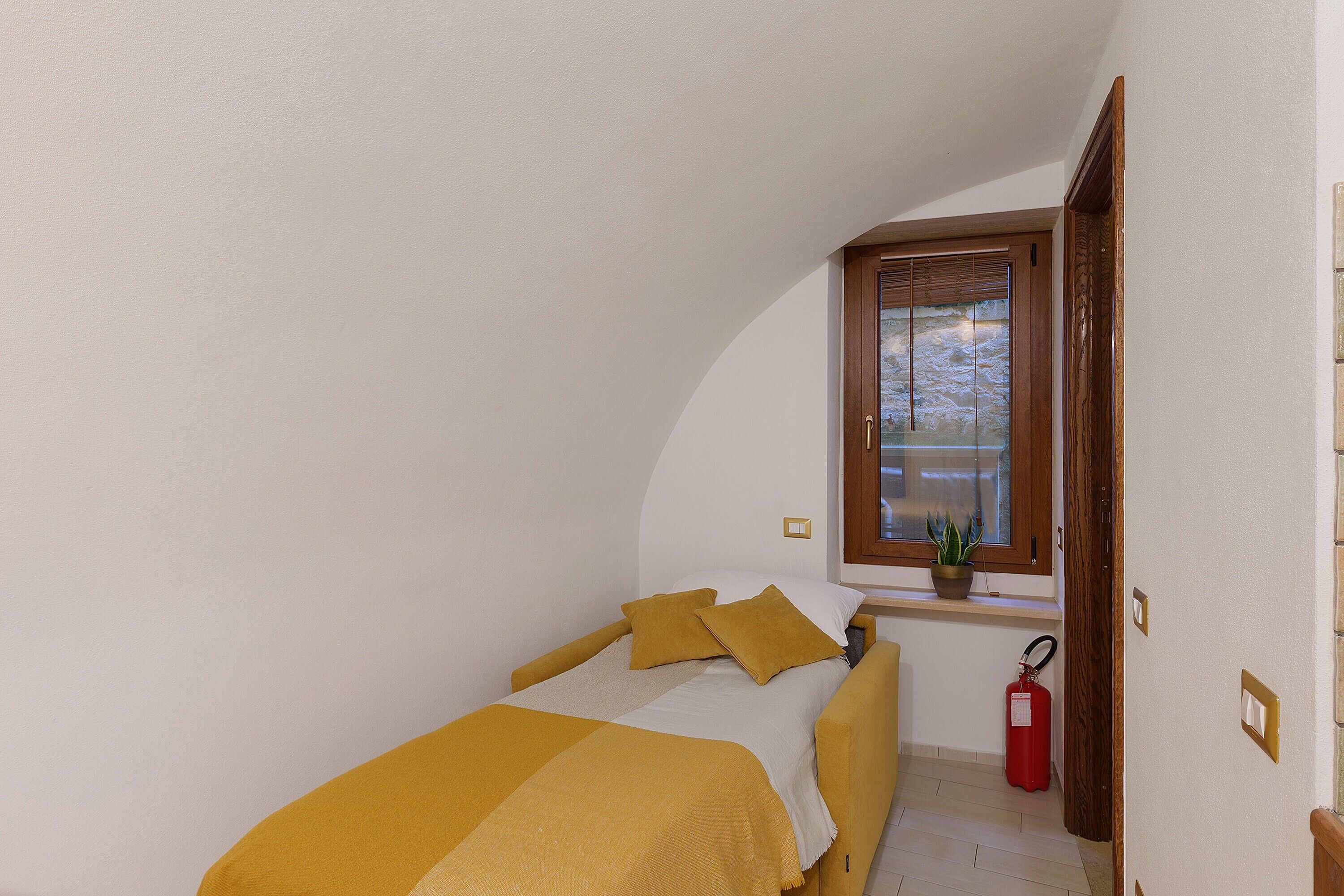 Een kleine slaapkamer met een eenpersoonsbed, een raam met uitzicht, een potplant en een brandblusser.