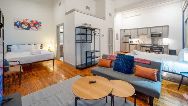 Espaço compacto para viver, com cama, sofá, duas mesinhas, pequeno espaço de cozinha e divisória de vidro.