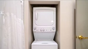 Uma lavanderia compacta e branca, com máquina de lavar e secadora embutidas, puxador dourado e cortina cobrindo parcialmente a porta.