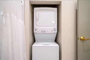 Un lavadero pequeño y blanco con lavadora y secadora empotradas, un picaporte dorado y una cortina que cubre parcialmente la puerta.