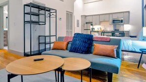 Uma sala de estar moderna com sofá, duas mesas redondas, roupeiro embutido e uma kitchenette ao fundo.