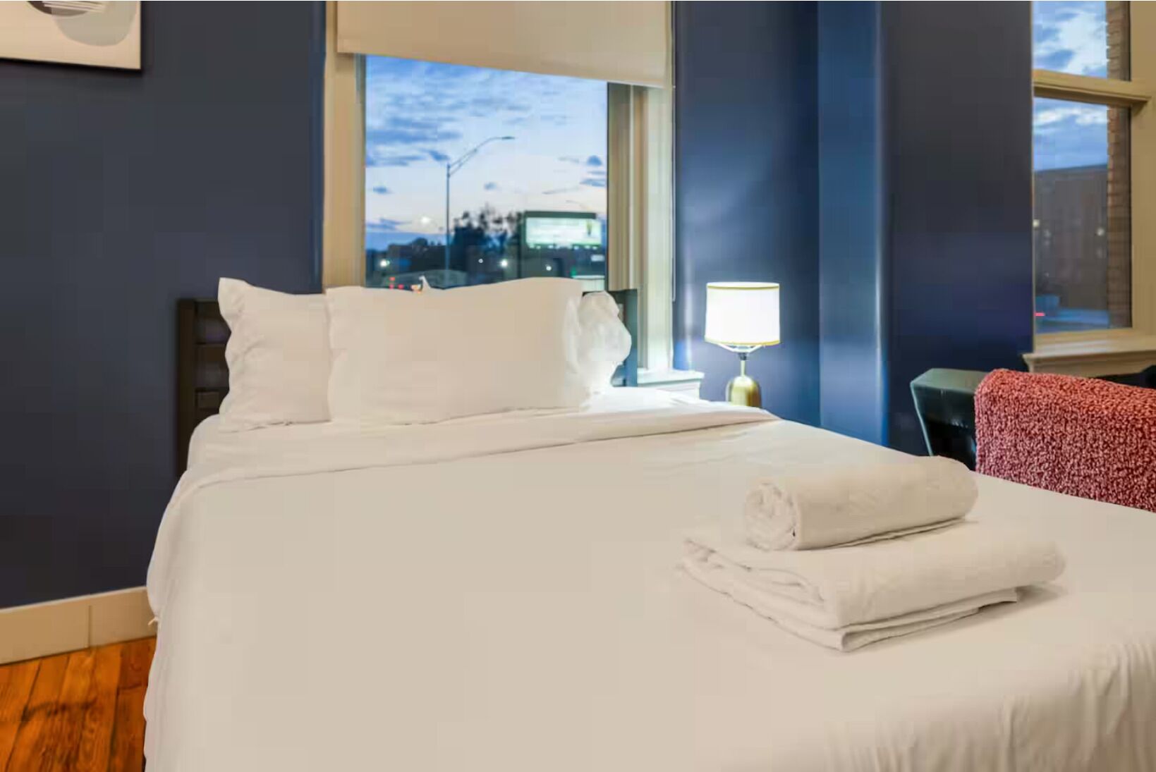 A hotel room with a neatly made bed, a lamp, and a view of the cityscape through the window.