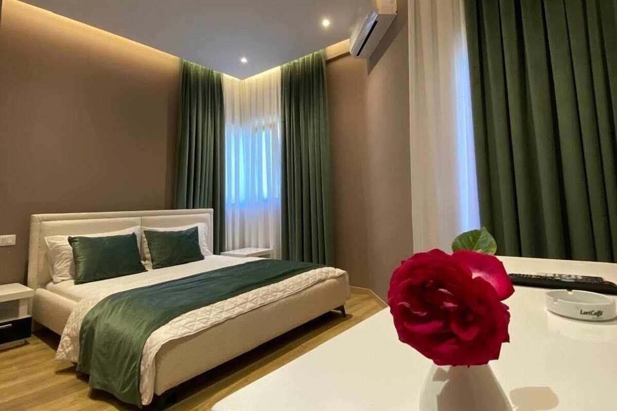 A modern hotel room with a bed, bedside tables, a vanity with a mirror, and a vase with a red rose.