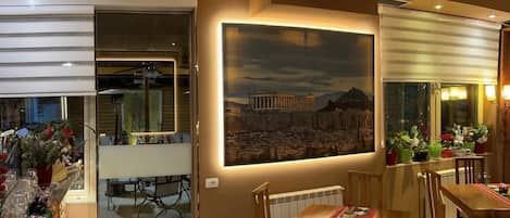 A dining area with a large painting of a historical building, wooden furniture, and potted plants.