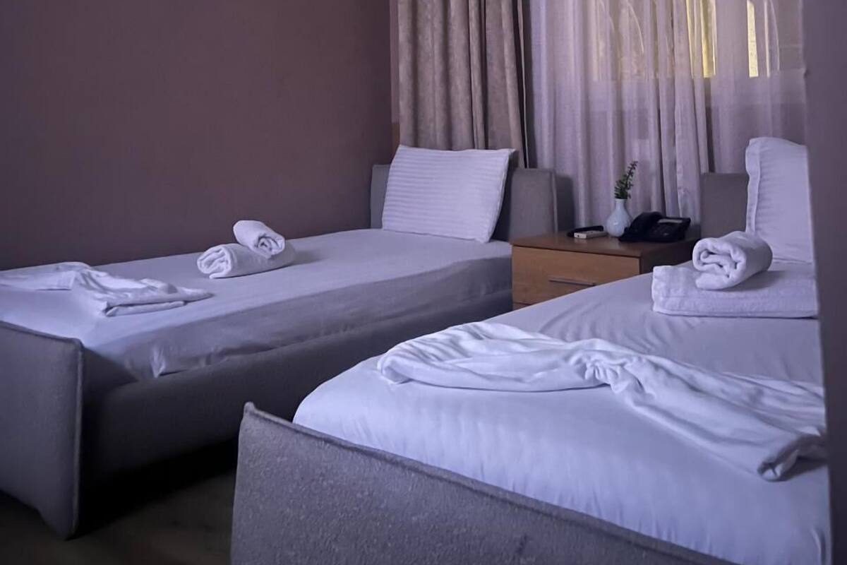 A hotel room with two single beds, a bedside table with a lamp, a wall-mounted air conditioner, and a window with curtains.