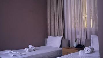 A hotel room with two single beds, a bedside table with a lamp, a wall-mounted air conditioner, and a window with curtains.