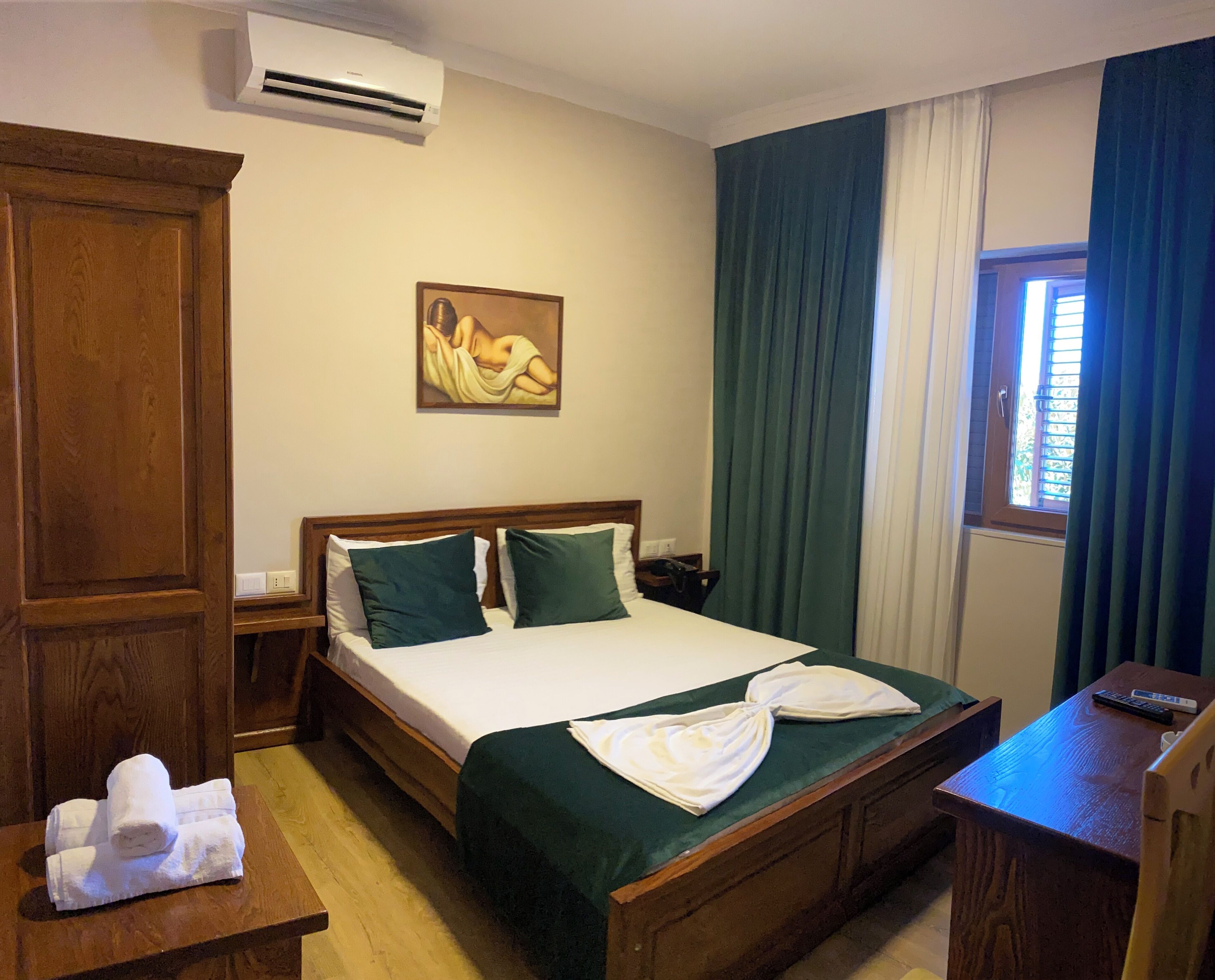 A hotel room with a wooden bed, green curtains, a painting on the wall, and a window with curtains.
