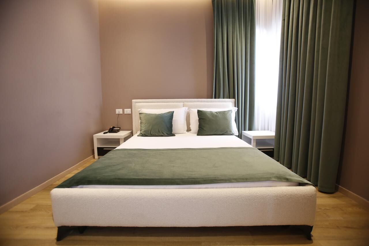 A neatly made bed with a green blanket, a white headboard, and two pillows. There is a bedside table with a phone and a lamp. The room has a window with curtains and a wooden floor.