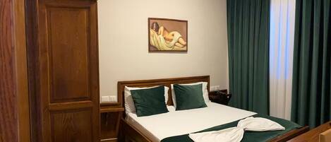 Una habitación de hotel con una cama de madera, ropa de cama verde, un cuadro en la pared, un aire acondicionado y una ventana con cortinas.