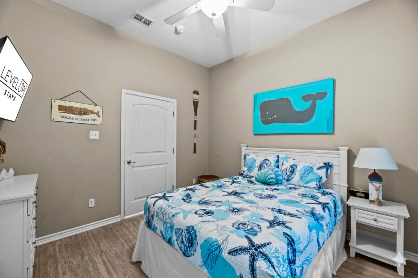 A bedroom with a bed, a nightstand, a lamp, a wall-mounted artwork of a whale, and a door.