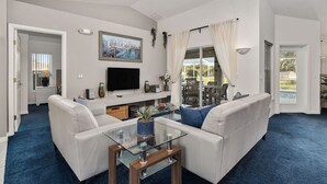 Een ruime woonkamer met een grote witte bank, een glazen salontafel en een flatscreen televisie aan de muur.