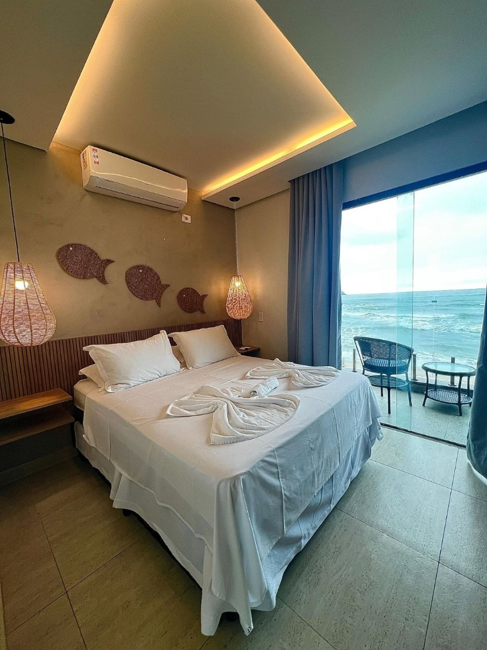 Habitación de hotel con una cama grande, un balcón con vistas al mar, una lámpara colgante y un aire acondicionado de pared.