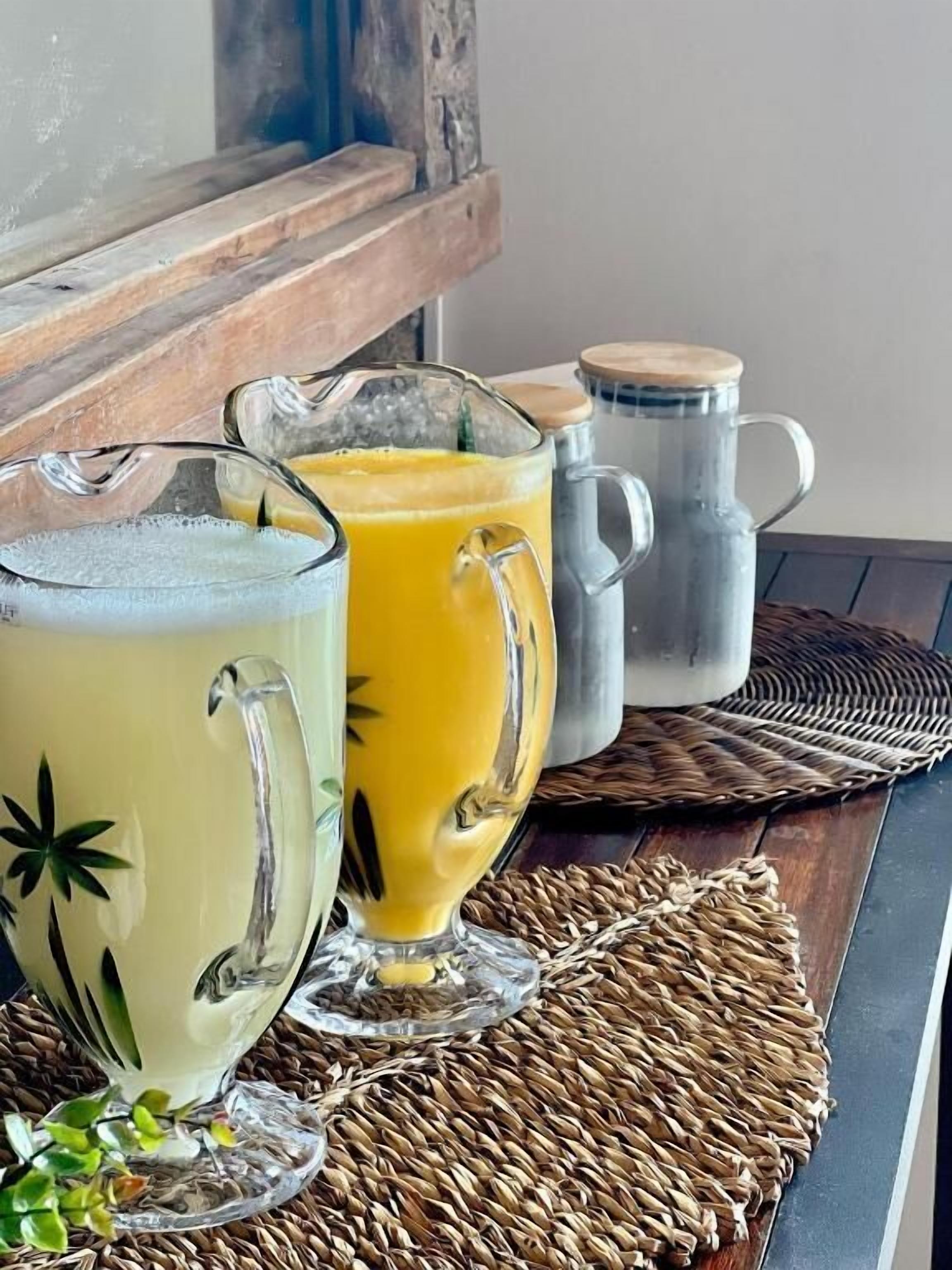 A wooden table with three glass pitchers of orange juice, a metal mug, and a woven placemat.