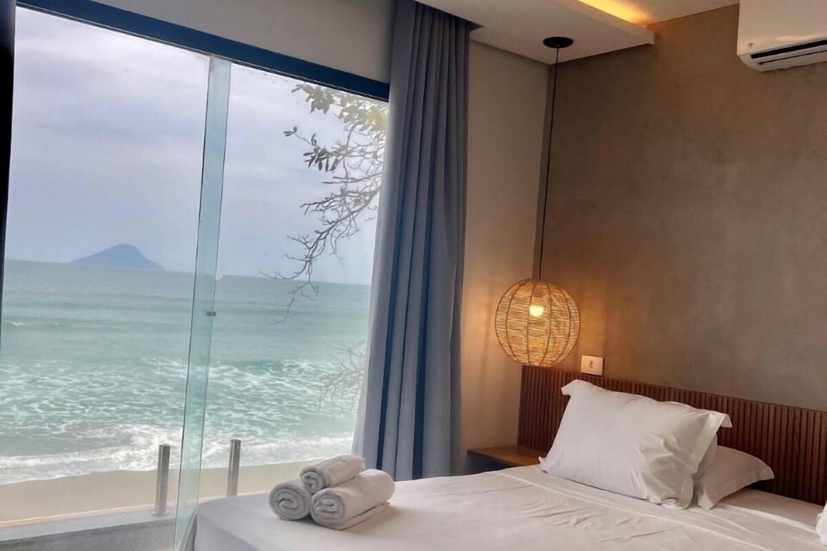 Une chambre d’hôtel avec un lit, une grande fenêtre donnant sur la mer, un climatiseur fixé au mur et une lampe de chevet.