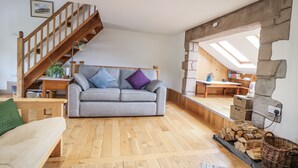 A spacious living area with a wooden staircase, a grey sofa with cushions, a wooden dining table, and a stone wall. - The Hayloft (Abergele)