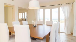 A dining area with a wooden table and white chairs, a pendant light, and a view of the sea through large windows. - Sa Figuera – Extraordinary Villa with Mountain Views, Wi-Fi, and Air Conditioning (Illes Balears)