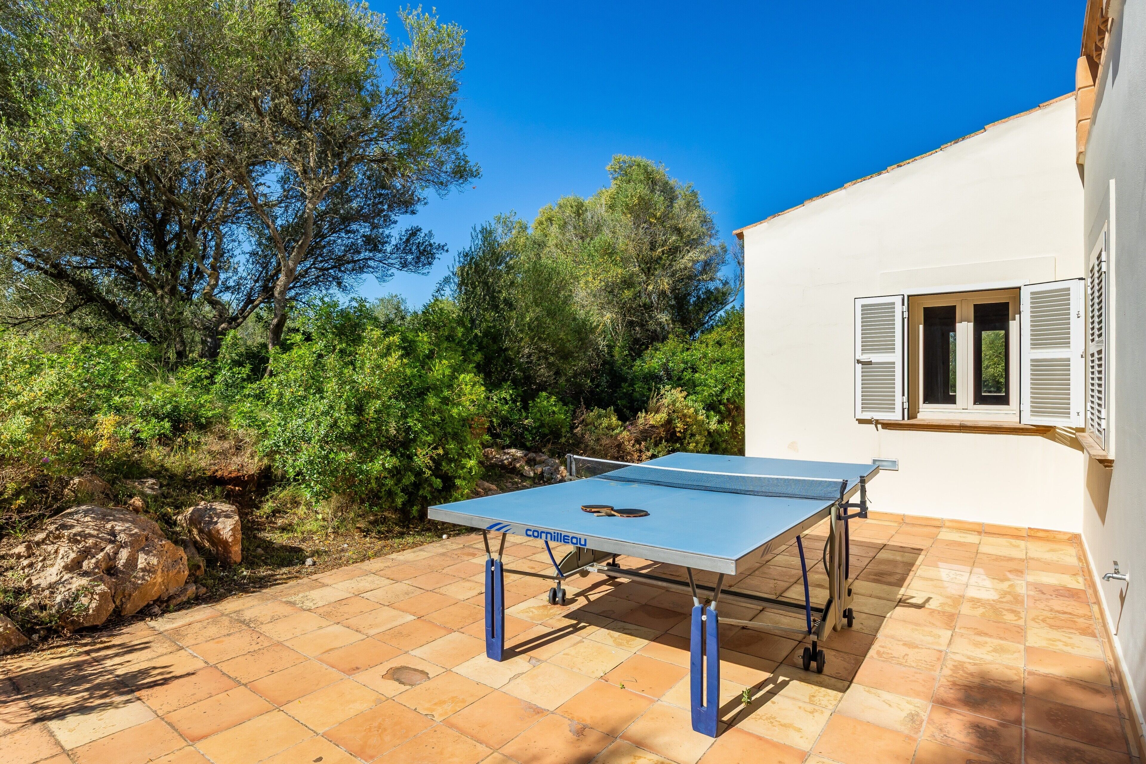 Une table de ping-pong sur une terrasse carrelée, avec un bâtiment blanc et des arbres en arrière-plan.