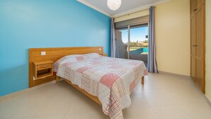 Quarto com cama, mesa de cabeceira, guarda-roupa de madeira, luz no teto e vista através de uma porta de vidro.