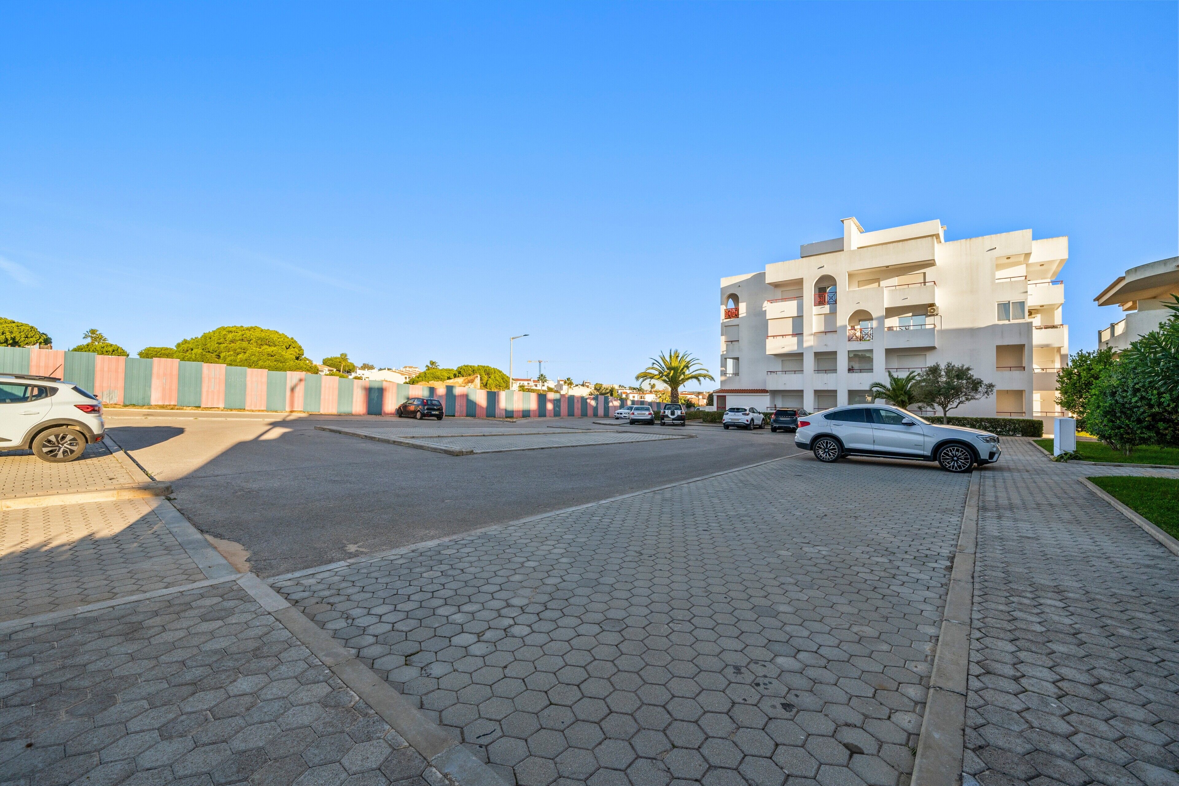 A paved parking area with cars, a multi-story building with balconies, and a clear blue sky.