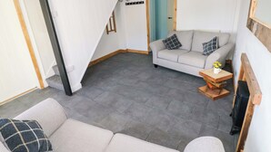 Una habitación con un sofá gris, una mesa de centro de madera, una mesita con un jarrón, una puerta azul y una escalera con paredes blancas y vigas negras.