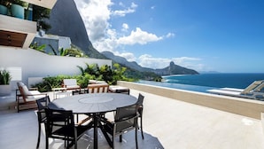 A modern outdoor dining area with a round table and chairs, overlooking a coastal view with mountains and the ocean.