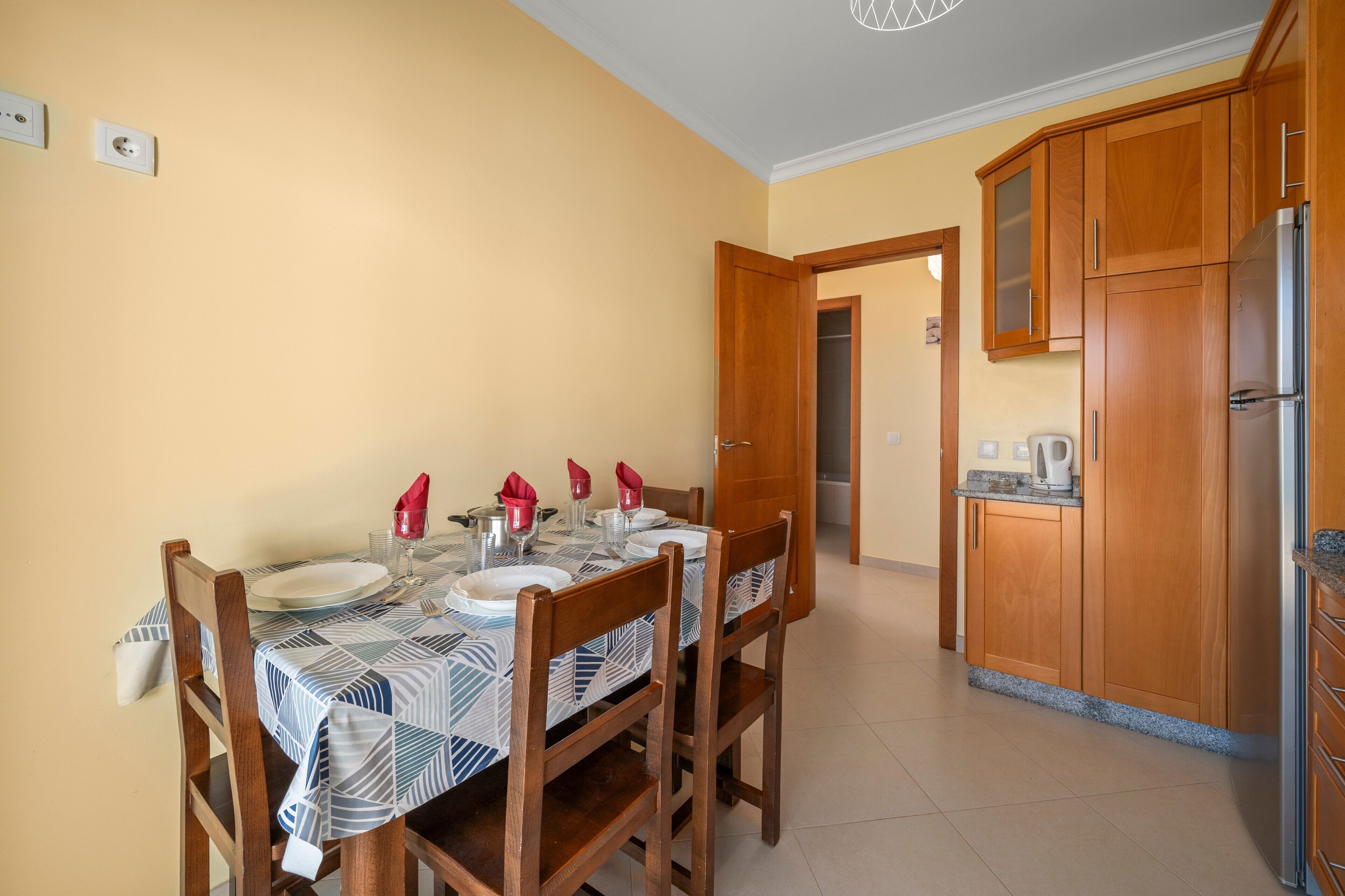 A dining area with a table set for four, wooden chairs, and a kitchen with wooden cabinets and a stainless steel refrigerator.