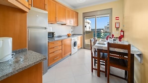 A compact kitchen with wooden cabinets, a built-in washing machine, and a dining area with a table and chairs. - 1-Bedroom Apartment with Shared Pool, Wi-Fi, and Air Conditioning (Armação de Pêra)