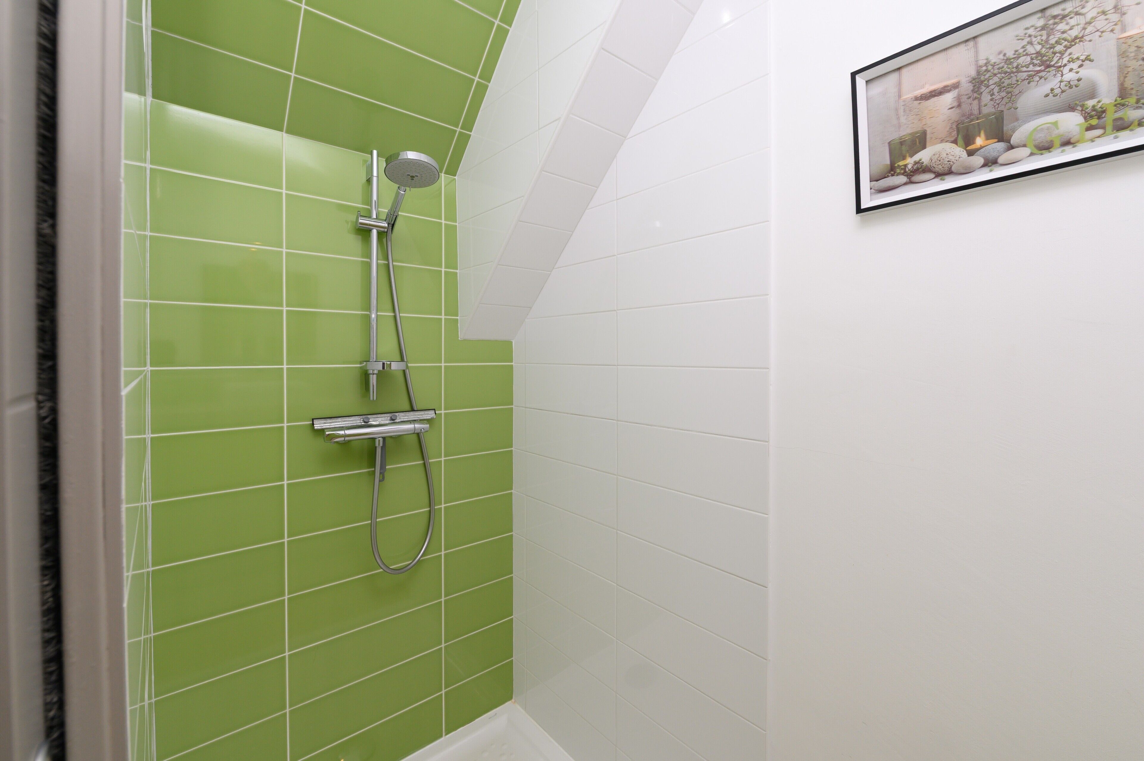 A bathroom with green and white tiled walls, a shower with a handheld showerhead, and a framed picture on the wall.