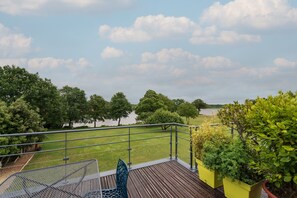 Un balcón con vista a un lago y árboles, una silla de metal y una terraza de madera.