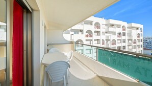 Un balcón con mesa y sillas, una cortina roja y vistas a un edificio blanco con balcones.