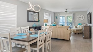 A dining area with a wooden table and white chairs, a ceiling fan, and a clock on the wall. - Waves & Rays - Stay, Play, and Soak Up the Day! (Horry County)