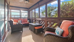 A covered outdoor seating area with wicker furniture, a ceiling fan, and large windows offering views of palm trees and a distant building. - Waves & Rays - Stay, Play, and Soak Up the Day! (Horry County)