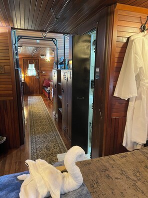 En hotellkorridor med träpanel på väggarna, en mönstrad matta, en bänk, en dörr med en handduksställning och en skymt av ett rum med en säng och ett fönster.