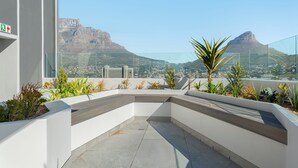 Une terrasse sur le toit offrant une vue sur une montagne et le paysage urbain, agrémentée de jardinières contenant diverses plantes et dotée d’une rambarde en verre.