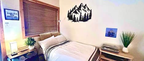 Une chambre avec un lit, une fenêtre avec des stores, un motif mural représentant des montagnes, une table de chevet avec une lampe et une petite étagère avec une plante.