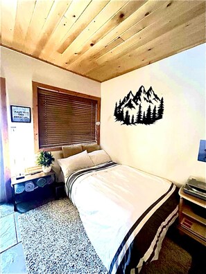 Une chambre avec un lit, une fenêtre avec des stores, un motif décoratif représentant des montagnes sur un mur, et un panneau fixé au mur.