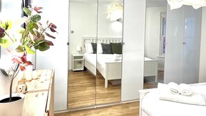 Ένα σύγχρονο δωμάτιο ξενοδοχείου με ξύλινο δάπεδο, ένα κρεβάτι με λευκά σεντόνια, ένα κομοδίνο, έναν καθρέφτη και ένα φυτό σε γλάστρα.