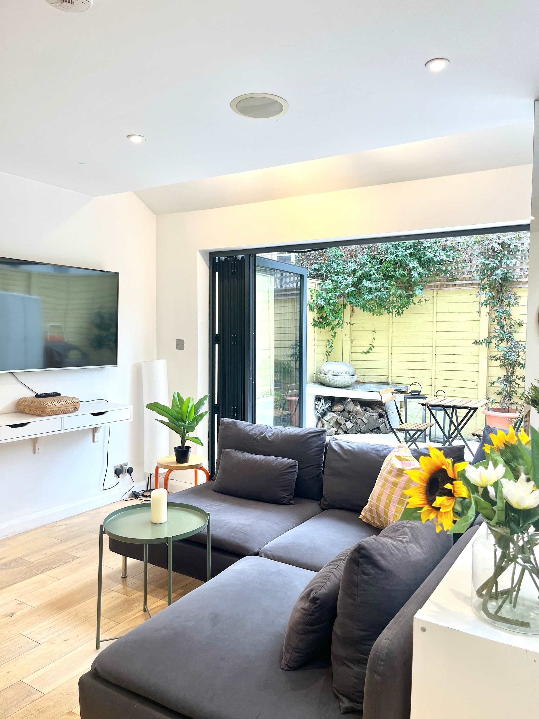 A modern living room with a grey sectional sofa, a small round table with a candle, and a large window offering a view of a garden and outdoor furniture.