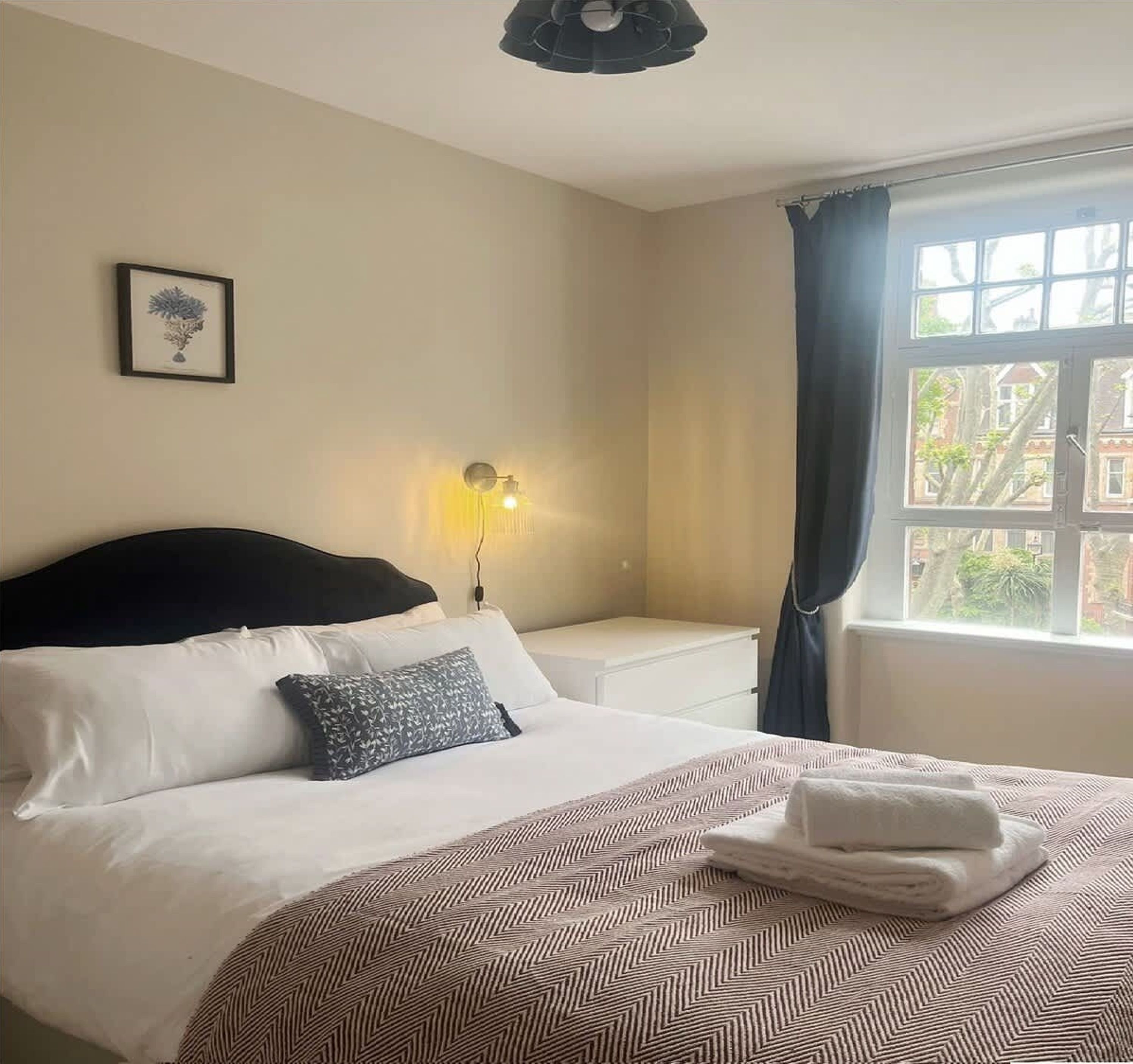 Une chambre d’hôtel avec un lit, une table de chevet, une lampe fixée au mur, une fenêtre avec des rideaux et un tableau encadré accroché au mur.