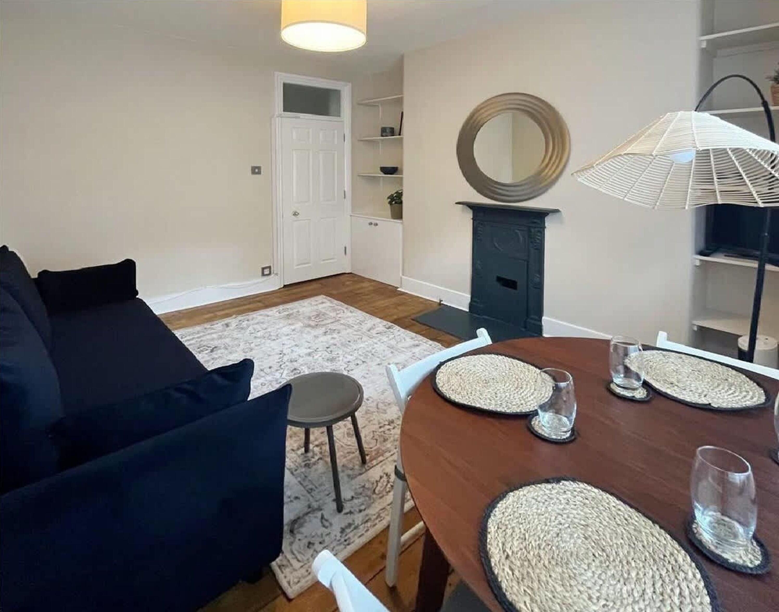 Ein Wohnzimmer mit einem dunkelgrauen Sofa, einem Esstisch mit gewebten Platzsets und einem großen Spiegel an der Wand.