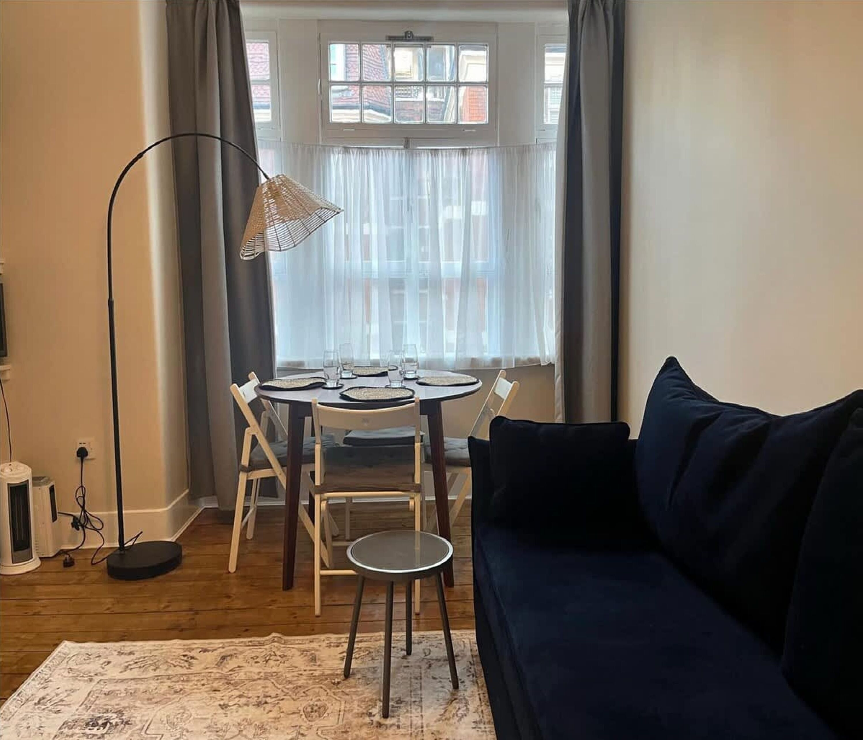 A living room with a dark blue sofa, a round dining table with chairs, a floor lamp, and a view through large windows.