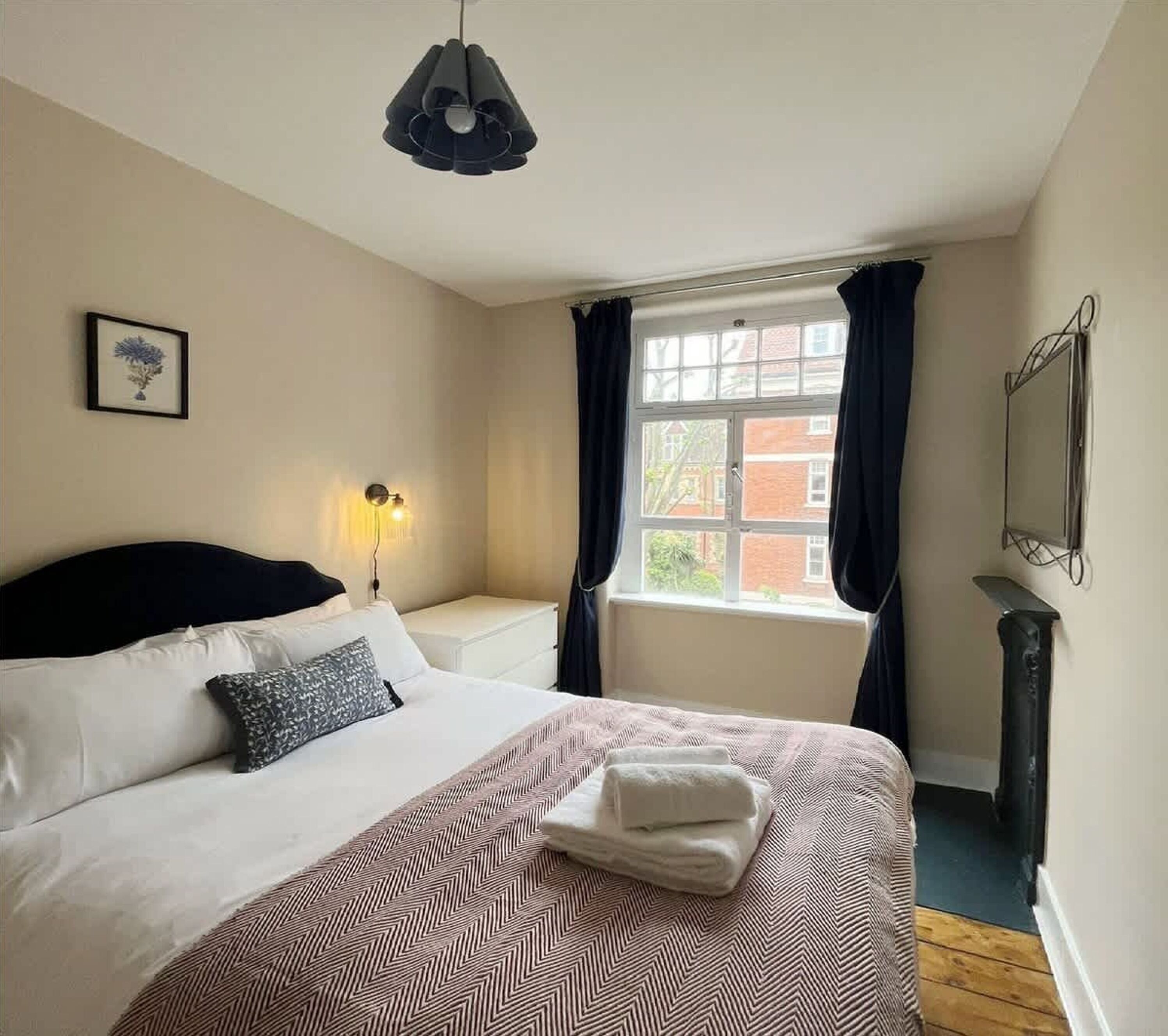 A bedroom with a bed, a bedside lamp, a window with curtains, and a framed picture on the wall.
