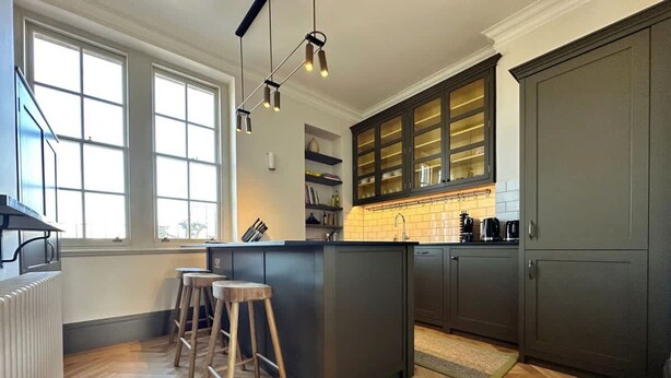 A modern kitchen with a central island, wooden stools, and a large window providing natural light. - Melody Apartament in Greater London (London)