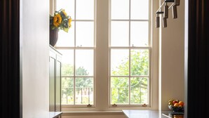 A kitchen with two windows, wooden stools, a radiator, and a fruit bowl on the counter. - Melody Apartament in Greater London (London)