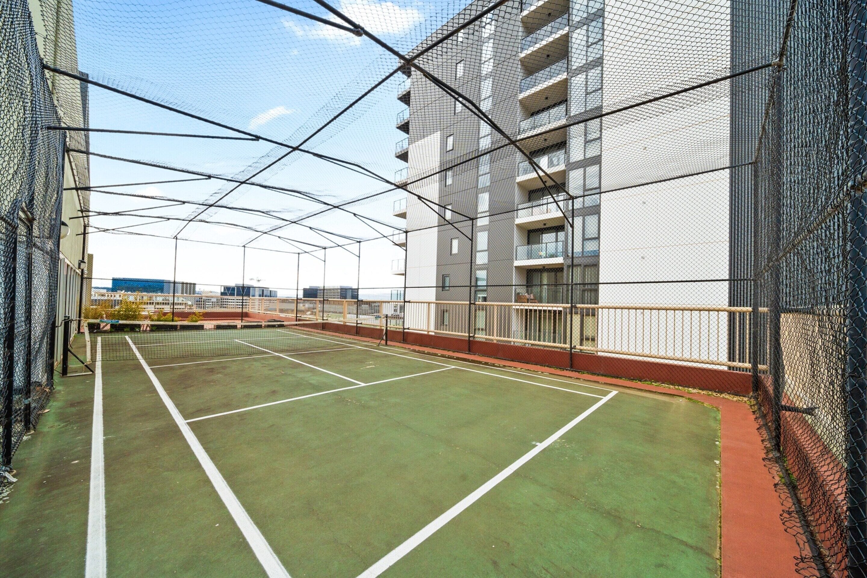 An indoor tennis court with a net, surrounded by chain-link fencing, and adjacent to a modern building with balconies.