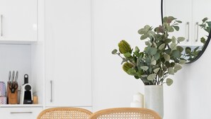 Een moderne keuken met witte kastjes, een glazen tafel, twee rieten stoelen, een vaas met groene bladgroenten, en een stapel boeken.