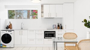 Een moderne keuken met witte kasten, een ingebouwde wasmachine, een magnetron en een glazen eettafel met rieten stoelen.