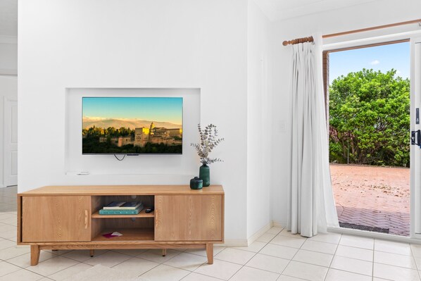 A modern living room with a flat-screen TV mounted on the wall, a wooden TV stand with books, and a large window with curtains. - Chic 1-bed Escape With Parking Bridgeman Downs (Brisbane)