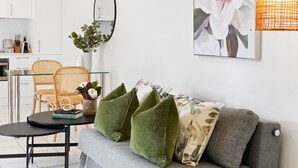 Een moderne woonkamer met een grijze bank, groene kussens, een ronde salontafel en een rieten stoel. Een groot schilderij met bloemen en een spiegel hangen aan de muur.